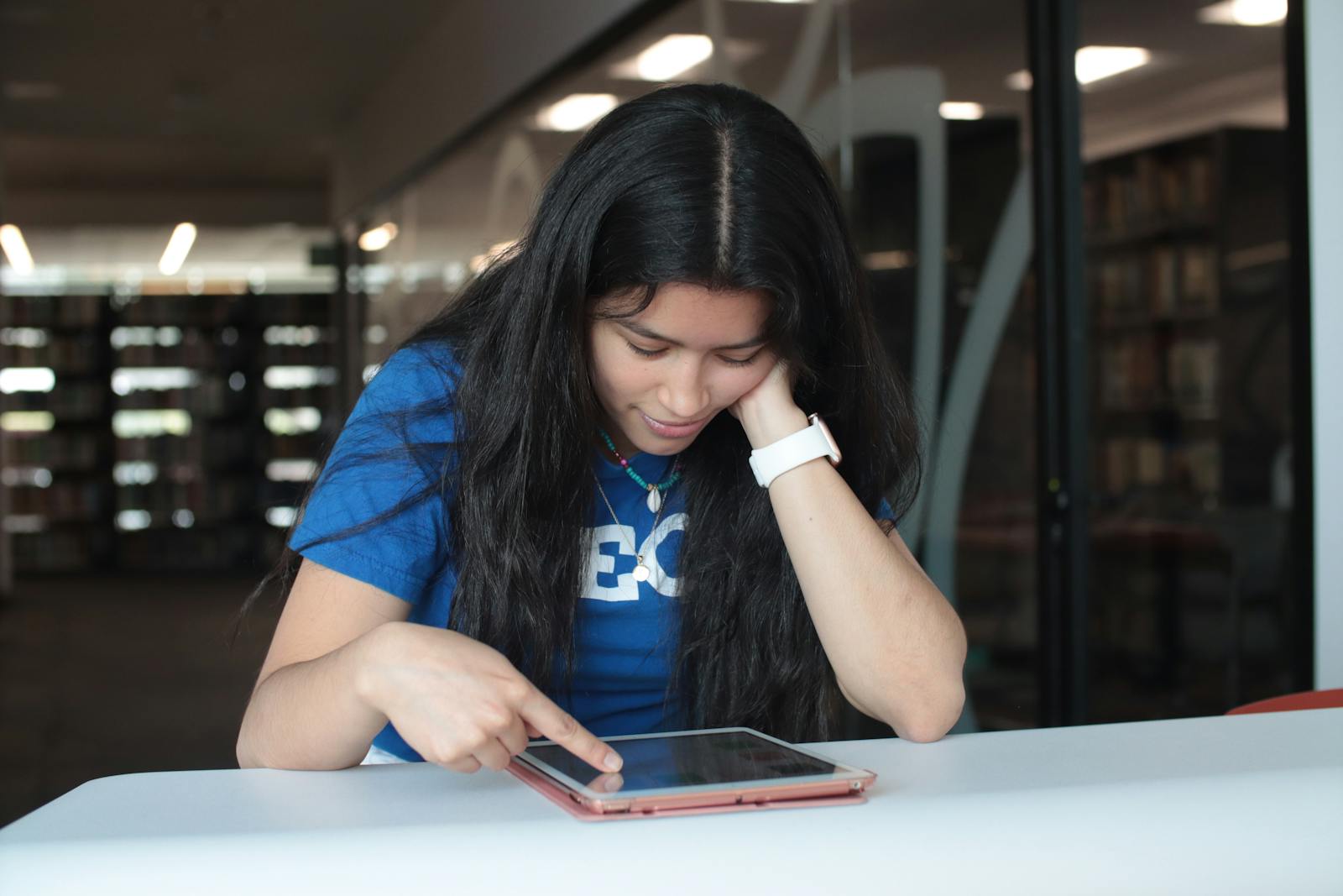 A young woman in a blue t-shirt is using a tablet at a library table. Focused and engaged in her task.