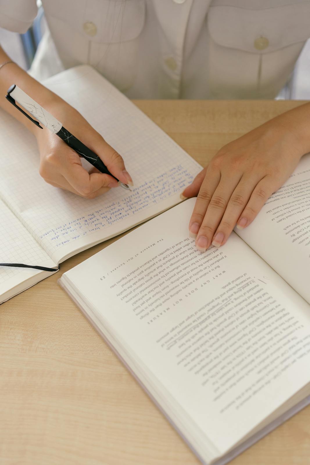 Adult writing notes in a notebook while reading a book indoors at a desk.