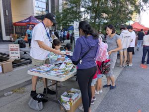 Literacy Now Burnaby table full of books surrounded by parents, children and BNH volunteers at the South Burnaby Summer Social 2025 event!