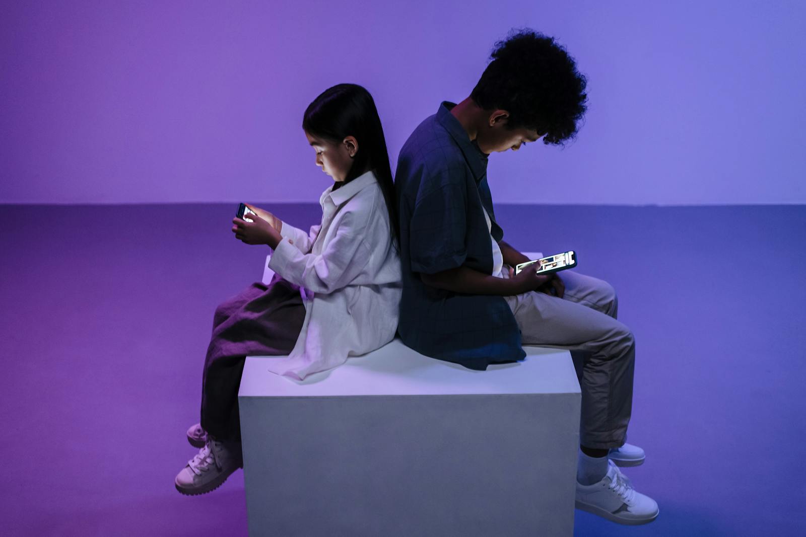 Two kids sitting back-to-back focused on smartphones, lit by moody lighting.